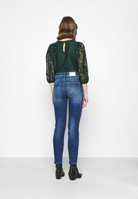 Top verde rendado com mangas bufantes e costas com recorte em forma de olho, combinado com jeans justos azuis e botas de tornozelo pretas de salto.