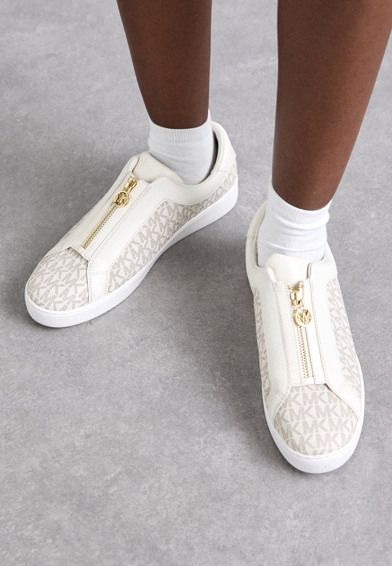 Baskets blanches avec motif « MK » beige et détail de fermeture éclair dorée, portées avec des chaussettes blanches aux chevilles sur un sol gris.