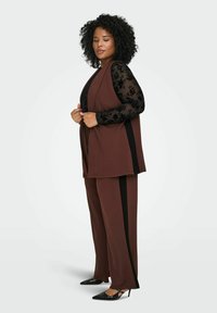 Žena s kovrčavom kosom koja nosi smeđi sako i hlače s crnim bočnim prugama, crnu cvjetnu čipkastu bluzu i crne štikle s oštrim vrhom.