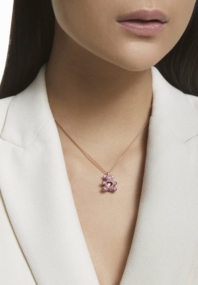 Un collier en or rose avec un pendentif en forme d'ours, orné de pierres précieuses roses et d'un design en cœur, reposant sur un blazer de couleur claire.