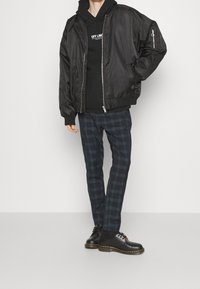 Giacca bomber nera con chiusura con zip, abbinata a un felpa nera con testo bianco e pantaloni a quadri blu navy. Scarpa nera con suola in gomma.