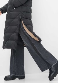 Persoon in een zwarte pufferjas met open zijrits, die donkere wijde jeans en zwarte chunky laarzen toont, tegen een effen witte achtergrond.