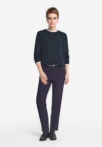 Granatowy sweter z białym dekoltem, zestawiony z dopasowanymi ciemnymi spodniami. Czarne buty o błyszczącej powierzchni. Model stoi z pewnością siebie.