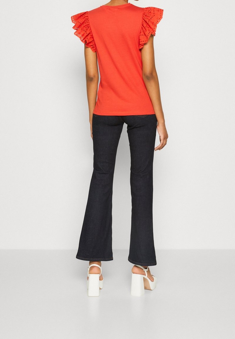 Czerwona koszulka z falbanowymi, ażurowymi rękawami, zestawiona z ciemnymi, rozkloszowanymi dżinsami i białymi sandałami na klockowym obcasie, widziana od tyłu.