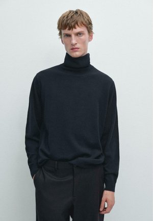 Pull noir à col roulé en matériau doux. Présente une coupe décontractée et des manches longues. Porté avec un pantalon noir ajusté, fond unicolore.