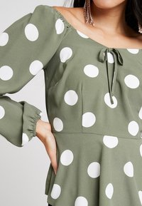 Blusa verde oliva con pois bianchi, design scollato sulla spalla, nodo al collo e polsini elastici per un look arricciato.