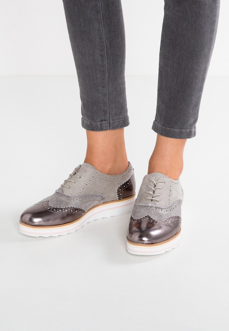 Chaussures en daim gris avec accents métalliques, arborant un design brogue, un bout rond et une semelle plateforme blanche. Surface texturée avec des détails de couture.