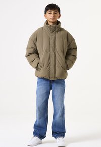 Chaqueta de plumas verde oliva con alto cuello, corte holgado y textura acolchada. Combinada con jeans de mezclilla azules y zapatillas blancas.