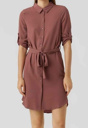 Vrouw draagt een dusty rose overhemdjurk met opgerolde mouwen en een strikceintuur in de taille tegen een effen achtergrond.