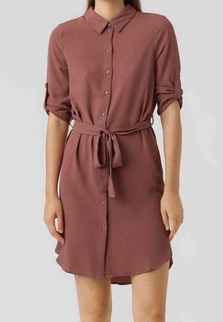 Femme portant une robe chemise rose poussière avec des manches retroussées et une ceinture nouée à la taille sur un fond uni.