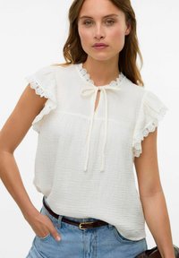 Vit texturerad blus med spetsdetaljer på ärmarnas kant och knytning vid halsen, bärs med högmidjade denimshorts.