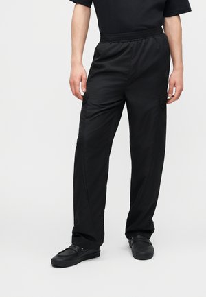 Άτομο που φοράει φαρδιά μαύρα παντελόνια cargo με ελαστική μέση και μαύρα παντόφλες, στέκεται σε απλό φόντο.