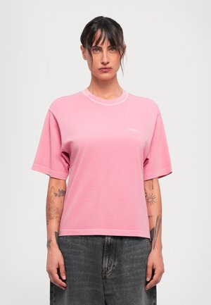 Frau mit dunklen, zurückgebundenen Haaren, die ein rosa T-Shirt mit dem Text "CLOSED" und dunkelgrauen Jeans trägt und vor einem schlichten weißen Hintergrund steht.