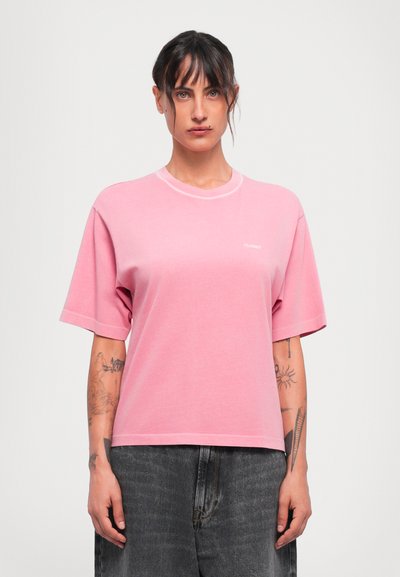 Mujer con el cabello oscuro recogido, vestida con una camiseta rosa que tiene el texto "CERRADO" y jeans gris oscuro, de pie frente a un fondo blanco liso.