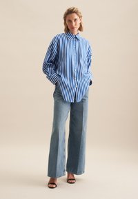 Blue and white striped oversized shirt with a button front and collar, paired with wide-leg light denim jeans and black sandals.