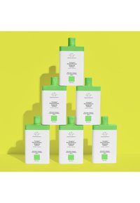 Six white shampoo bottles with green caps stacked in a pyramid against a bright yellow background. Labels include product name and details.