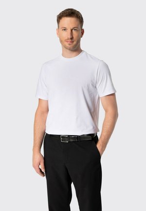 Hombre de pie con una mano en el bolsillo vistiendo una camiseta blanca lisa, pantalones negros y un cinturón negro sobre un fondo claro.