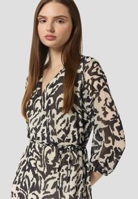 Przezroczysta, wzorzysta sukienka w czerni i kremie z dekoltem w serek, długimi rękawami i paskiem w talii. Charakteryzuje się luźnym krojem i lekką fakturą.