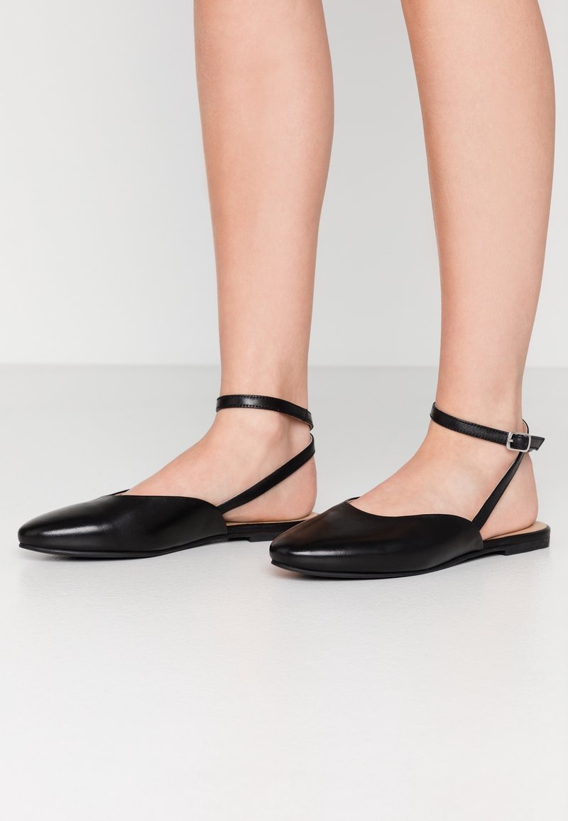 Chaussures plates en cuir noir avec brides à la cheville portées pieds nus sur un fond blanc.