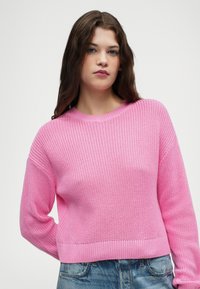 Roza pletena pulover s teksturirano površino, okroglim izrezom in rebrastimi manšetami ter robom, v kombinaciji z denim hlačami.