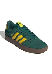 Zelené semišové tenisky se třemi žlutými pruhy, polstrovaným límcem a hnědou gumovou podrážkou. Zaoblená špička a zavazování vpředu.