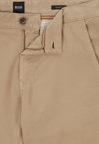 Béžové kalhoty se zúženým střihem vyrobené z hladké bavlněné tkaniny. Mají zip na předním díle, zapínání na knoflík a boční kapsu s zesíleným šitím.