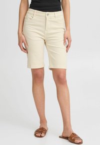 Shorts en denim beige avec une coupe droite, des revers, des poches avant et une fermeture à bouton. Associés à des sandales brunes à brides.