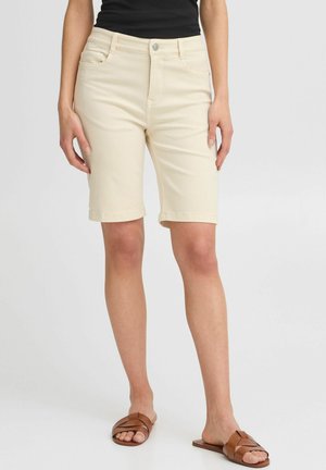 Shorts en denim beige avec une coupe droite, des revers, des poches avant et une fermeture à bouton. Associés à des sandales brunes à brides.