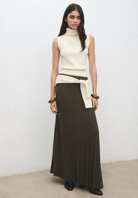 Top crem fără mâneci cu guler înalt, combinat cu o fustă lungă, verde închis, cu dungi, și o curea crem. Cizme simple negre până la gleznă.