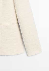 Giacca beige chiaro in tessuto strutturato, caratterizzata da una silhouette squadrata e un davanti leggermente aperto. Maniche lunghe con orli grezzi.