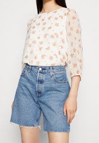 Blusa floreale bianca con stampa rosa e verde, maniche lunghe trasparenti. Abbinata a pantaloni shorts in denim blu a vita alta con orlo sfrangiato.
