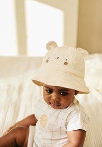 Kremna vedrovka s medvedjimi ušesi in obraznimi detajli, mehka tekstura. Otrok nosi bel pajac s karo vzorcem in grafiko sonca.