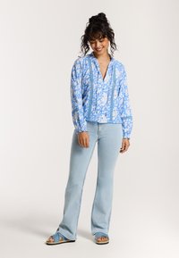 Blouse bleu à motifs floraux avec des manches longues et un col en V, associée à un jean flare bleu clair et des sandales bleues, avec un tissu texturé.
