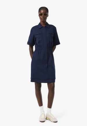 Femme portant une robe courte bleu marine à manches courtes avec des poches avant, des chaussettes blanches, des baskets roses et des lunettes de soleil, debout les mains derrière le dos.