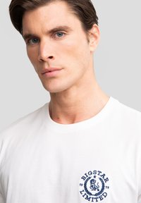 Hvid bomulds T-shirt med rund hals, prydet med et mørkeblåt broderet logo af en løve inden for et cirkulært design på brystet.
