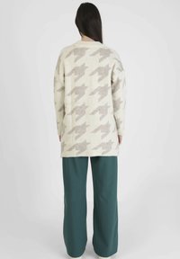 Femme aux longs cheveux foncés portant un pull surdimensionné beige et gris à motif pied-de-poule et un pantalon ample vert foncé, debout de dos.