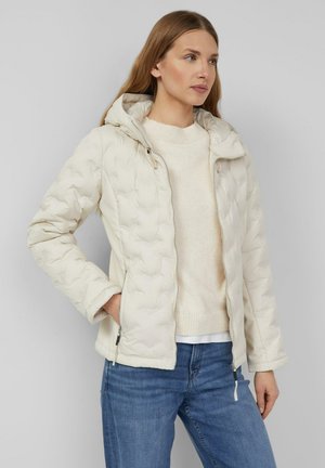 Femme portant une veste matelassée à capuche de couleur crème par-dessus un pull léger avec un jean bleu, debout avec une main dans la poche de la veste, regardant sur le côté.