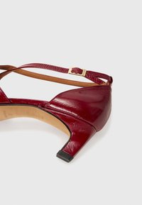 Chaussure à talon haut en cuir bordeaux avec un bout pointu, une bride de cheville et une boucle dorée, présentant une finition texturée et un petit talon noir.