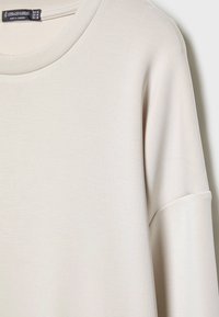 Jasnobeżowy sweter z okrągłym dekoltem, wykonany z gładkiego materiału, z krótkimi rękawami i luźnym krojem. Metka widoczna na dekolcie.