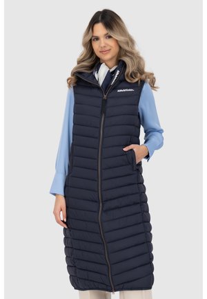 Frau trägt eine lange, marineblaue, gesteppte ärmellose Weste mit Reißverschluss vorne und eine hellblaue Langarmbluse, steht mit einer Hand in der Tasche.