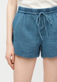 Shorts bleu texturé à cordon portés avec un haut blanc, montrant la taille, les hanches et un bras pendant détendu le long du corps.