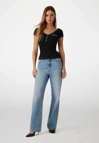 T-shirt preto justo com mangas curtas e detalhes em botão, combinado com jeans de pernas largas azul-claro e sapatos de salto alto pretos.