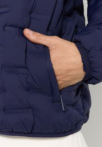 Granatowa kurtka puchowa z zamykaną na zamek kieszenią, teksturowaną tkaniną i przylegającym krojem. Materiał wydaje się być wodoodporny i lekko błyszczący.