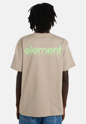 Person mit dunklen Dreadlocks, die ein beiges T-Shirt mit grünem Schriftzug "element skateboard co. worldwide" auf dem Rücken trägt, kombiniert mit blauer Jeans.
