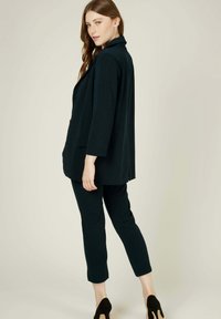 Blazer noir et pantalon ajusté assorti en tissu lisse. Le blazer a une coupe décontractée avec une poche avant unique et une ouverture frontale. Les talons sont noirs.