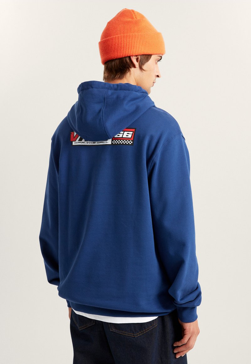 Blauwe hoodie met een ontspannen pasvorm, voorzien van een rugafbeelding met rode en zwarte accenten en een geruit patroon. Gecombineerd met een feloranje beanie.