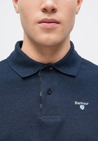 Ναυτικό μπλε πόλο μπλούζα από υφασμένο βαμβάκι, με κλασικό γιακά, δύο κουμπιά και κεντημένο λογότυπο στο στήθος.