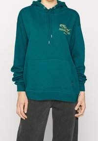 Osoba ubrana w miętowy hoodie z żółtym napisem "Athletic club" na piersi oraz ciemnoszare dżinsy, stojąca z rękami swobodnie opuszczonymi wzdłuż boków.