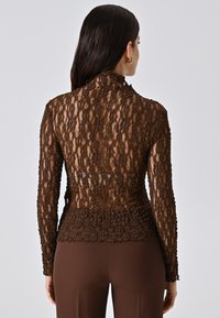 Top lungo a maniche profonde in pizzo marrone con scollatura alta. Presenta un motivo floreale testurizzato con retro trasparente. Abbinato a pantaloni marroni aderenti.