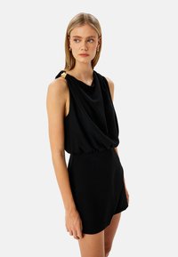 Jumpsuit negro sin mangas con frente drapeado, cintura fruncida y acento dorado en el hombro. Tejido suave con un ajuste holgado y diseño de pantalones cortos.